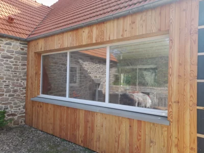 Pose d’une large baie vitrée aluminium sur extension bois avec bardage, apportant luminosité et modernité à une rénovation de maison en pierre.